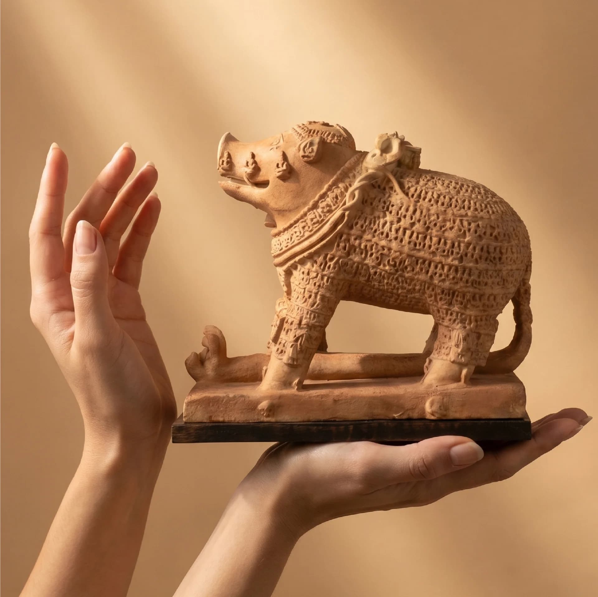 A pair of hands gently cradles an intricately carved terracotta-style Adi Varaha Sculpture, showcasing the detailed boar-headed deity replica.