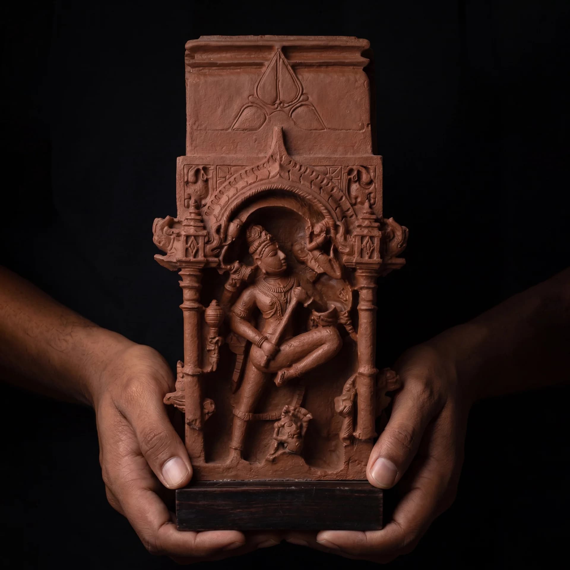 Two hands hold a Gajasura Vadha Sculpture, a stone composite replica showing intricate temple relief carvings of Shiva.