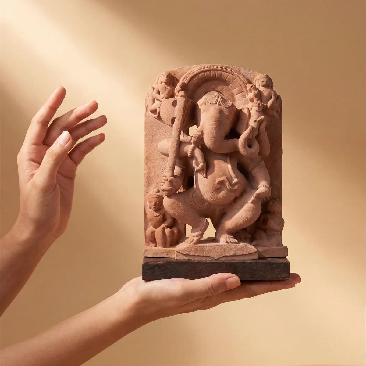 A pair of hands gently cradles a handcrafted Ganesha Sculpture, a sandstone-style replica featuring intricate temple art details.
