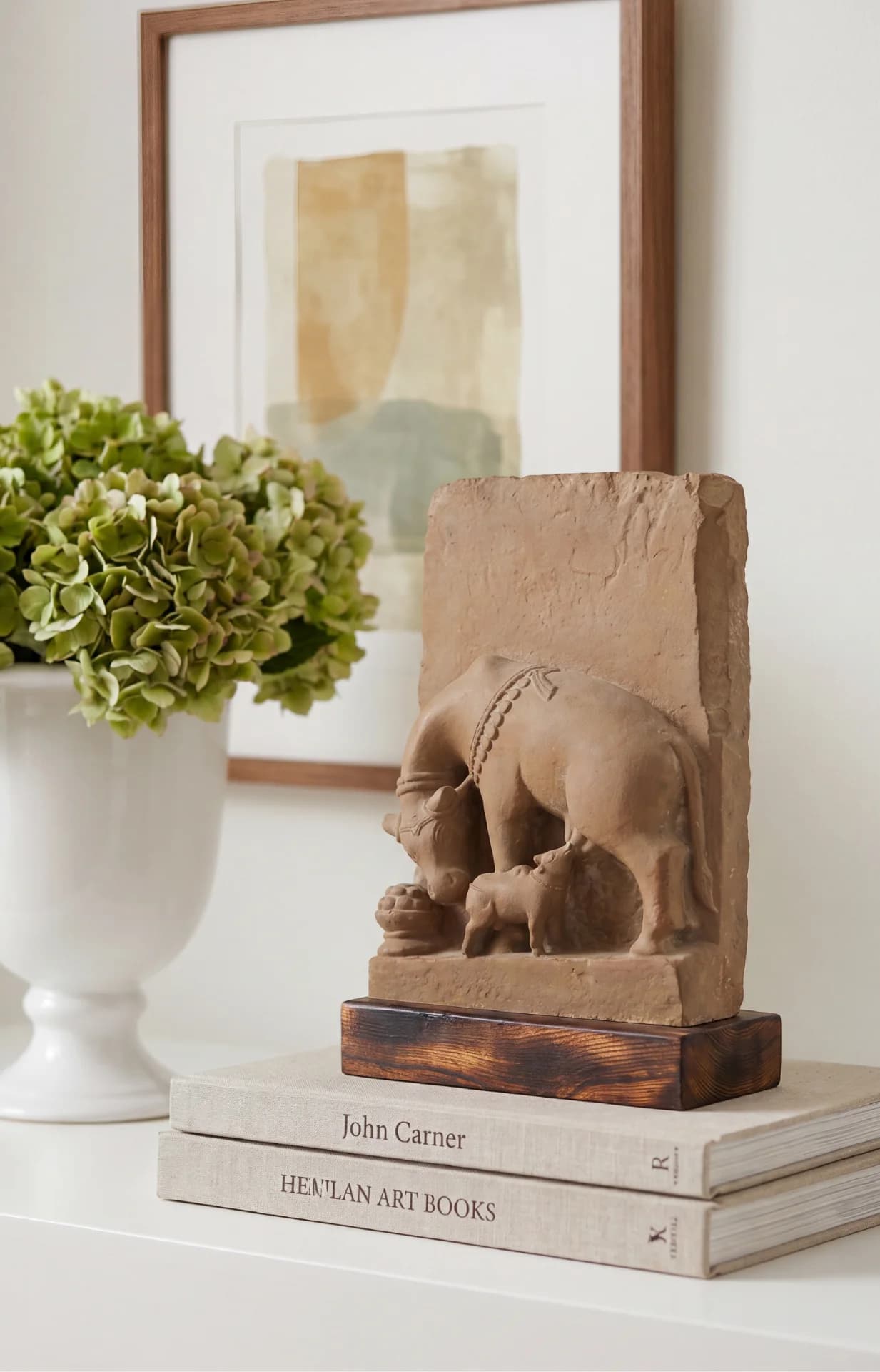 A stone composite Kamdhenu Sculpture displayed on books next to a white vase with green flowers.