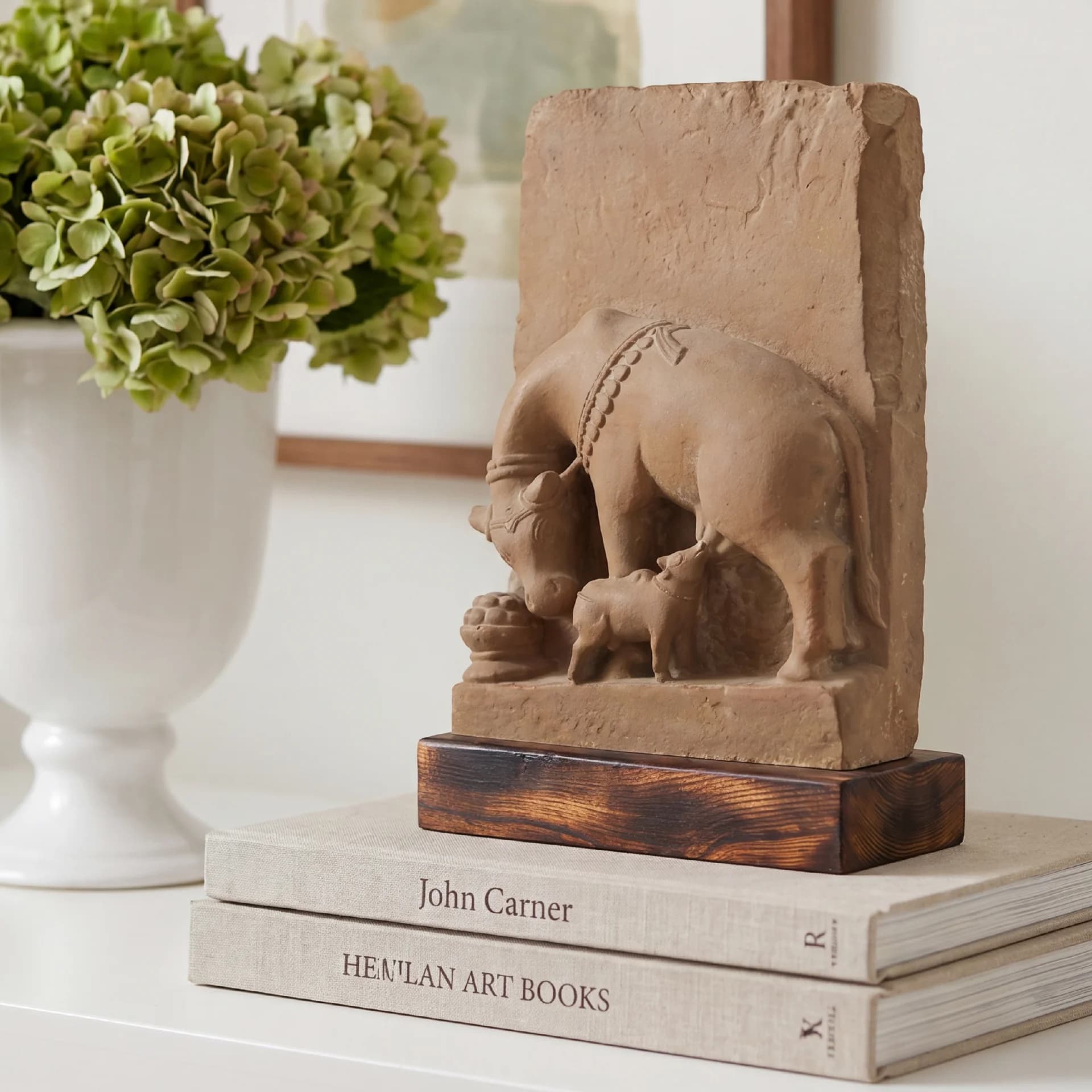 A stone composite Kamdhenu Sculpture displayed on a stack of books next to a white vase with green flowers.