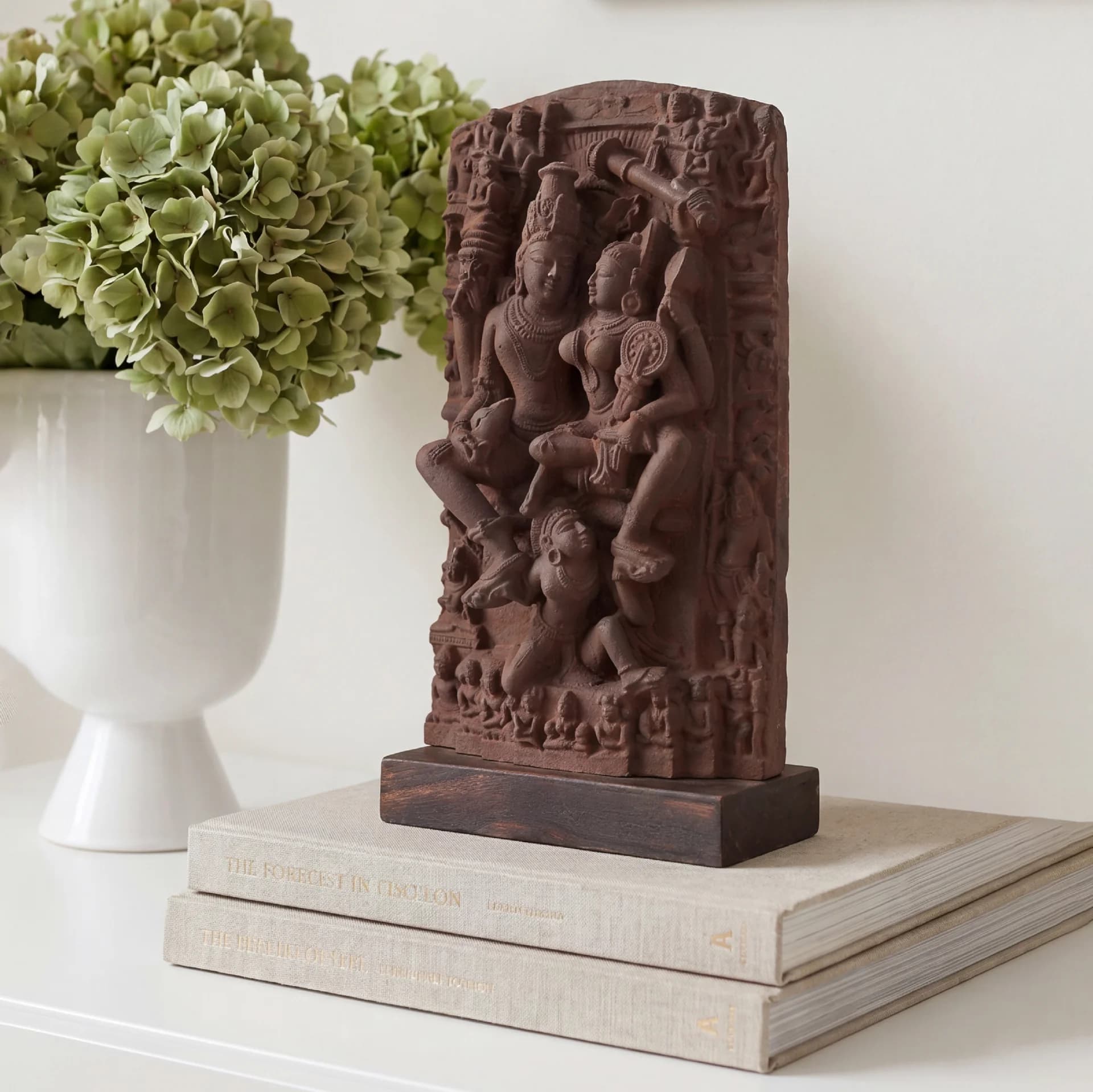 A detailed terracotta-style Lakshmi Narayan Sculpture displayed on stacked books next to a white vase with green flowers.