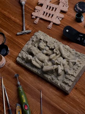 A high-angle view of a sandstone relief sculpture being crafted on a wooden workbench surrounded by various carving tools.