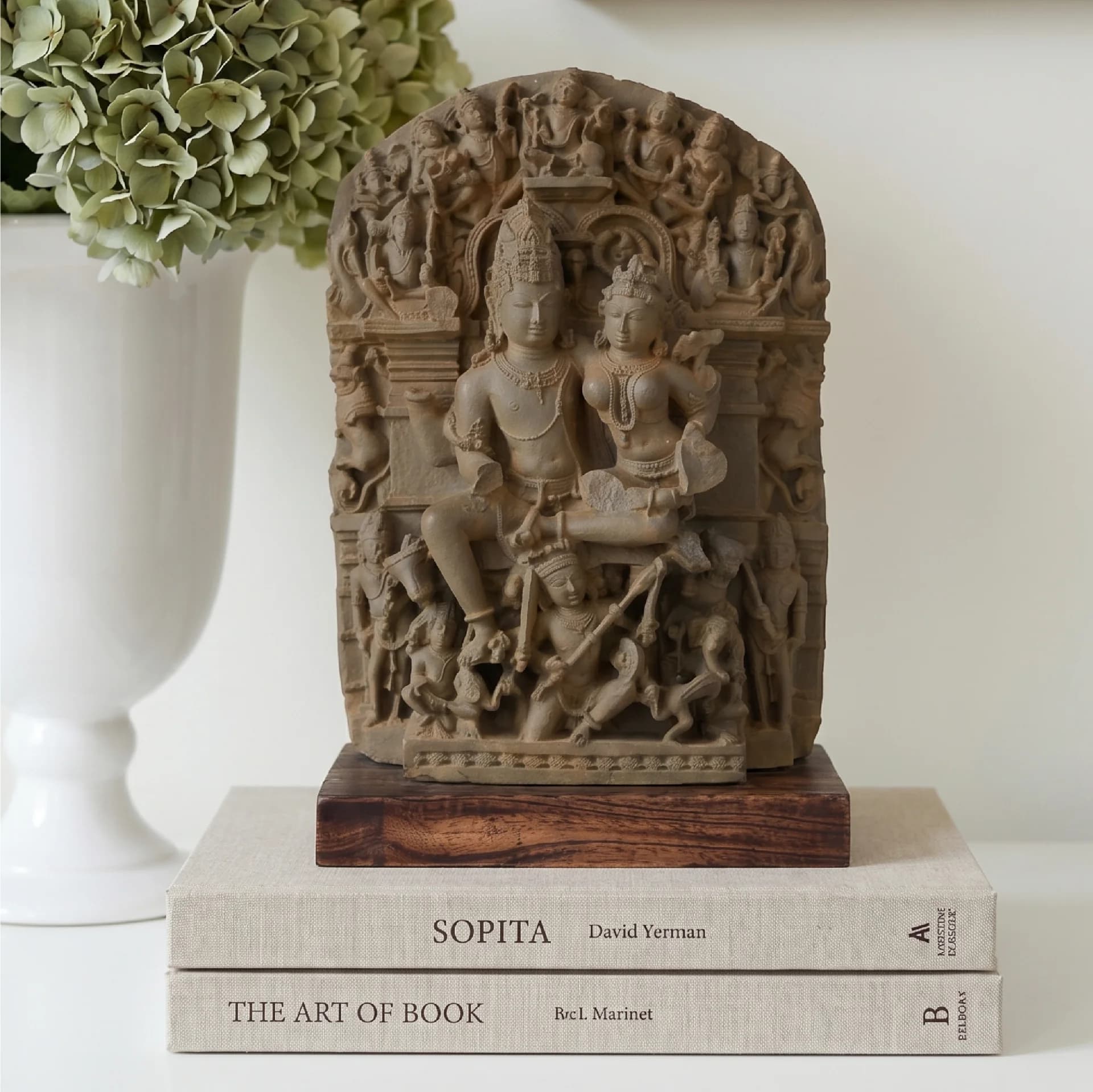 A front-facing view of the Ravananugraha Sculpture, a stone composite replica featuring intricate carvings, displayed on books with a floral arrangement.
