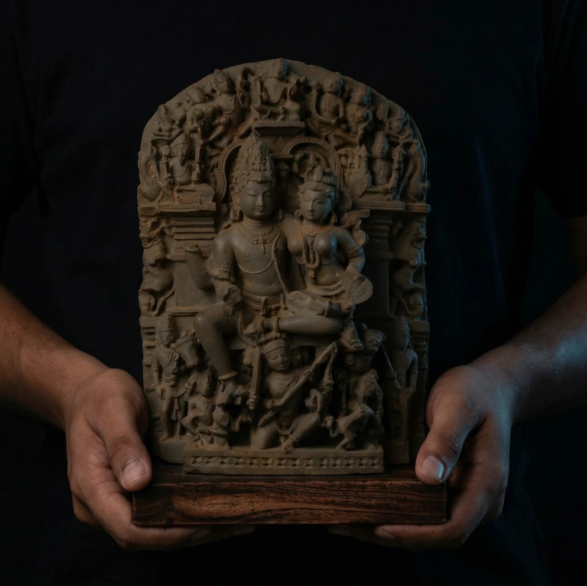 A pair of hands holds the Ravananugraha Sculpture, a detailed stone composite replica featuring Shiva and Parvati against a dark background.