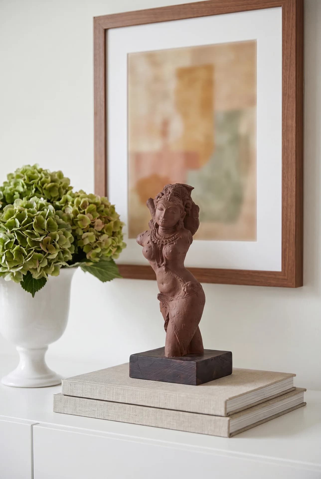 A stone composite Salabhanjika Sculpture displayed on stacked books next to a vase of green hydrangeas in a home setting.