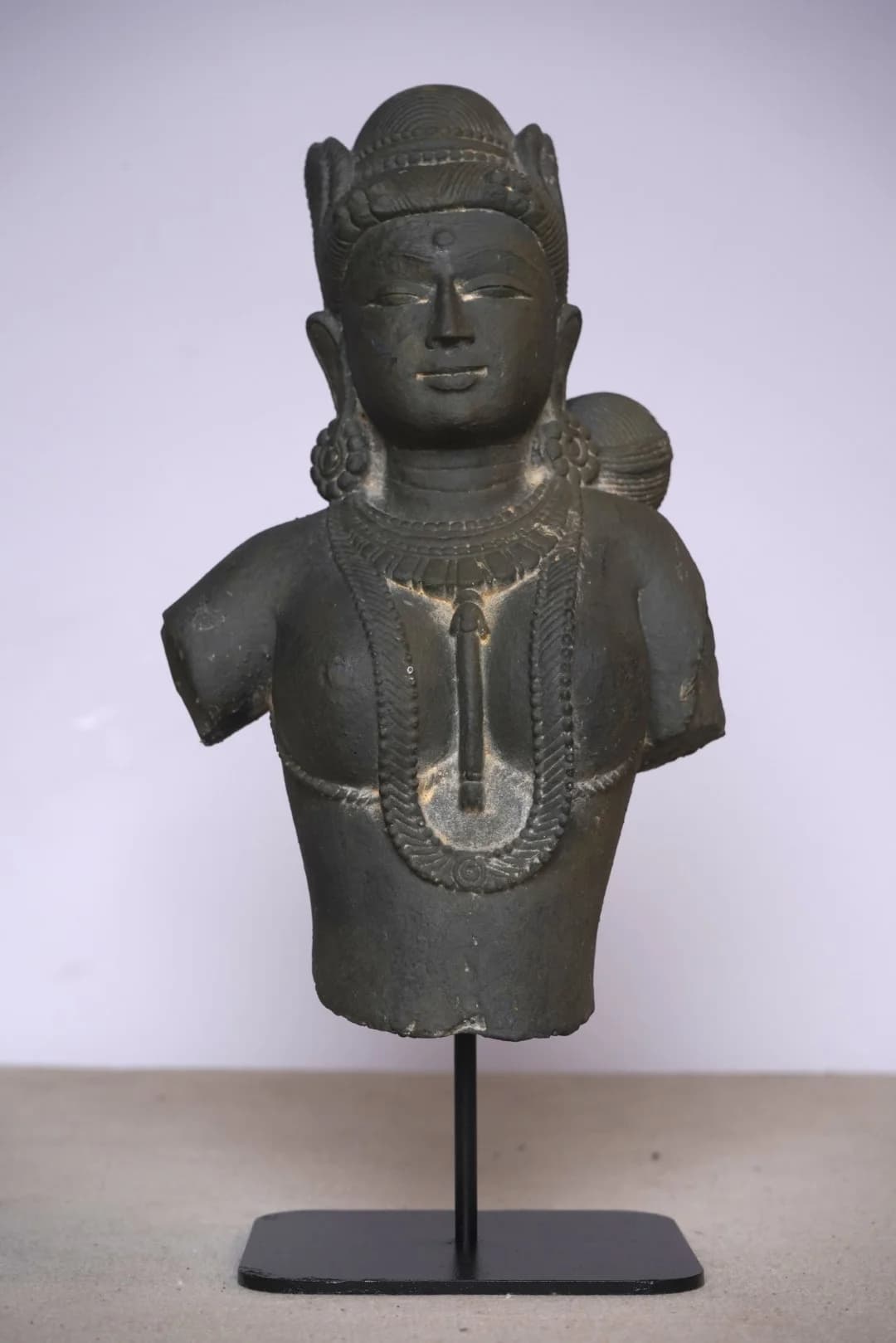 A front-facing view of the Test-BOM stone bust, featuring intricate carved jewelry and a serene expression on a display stand.