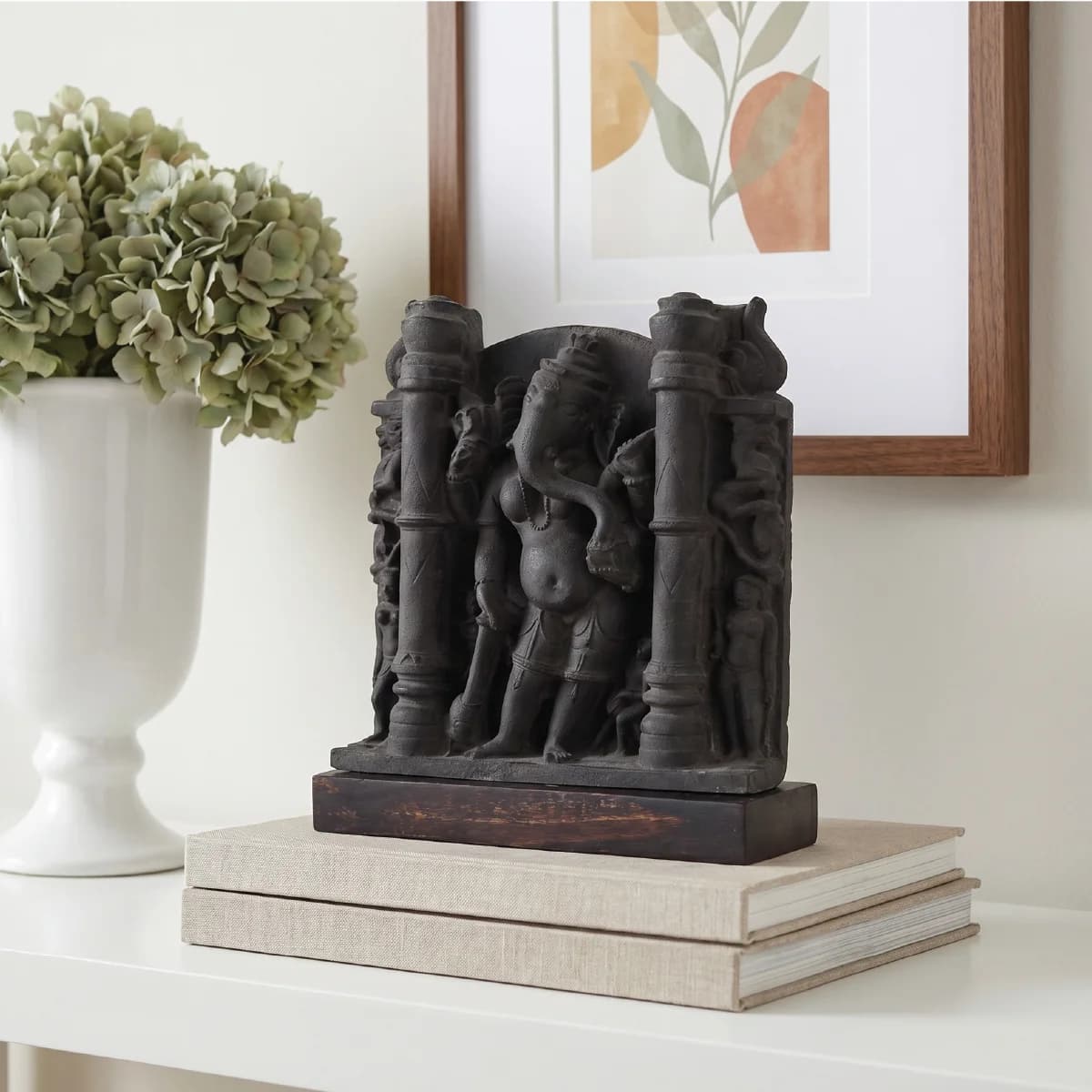 A dark stone Vinayaki Sculpture replica displayed on stacked books next to a white vase and framed art.