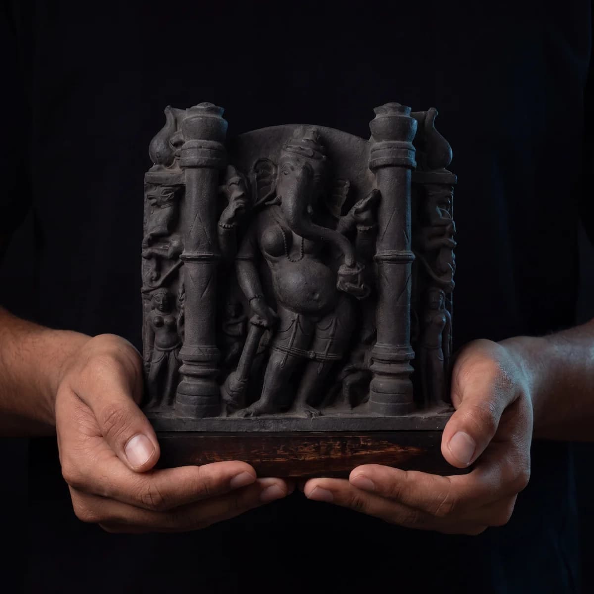 A pair of hands holds a dark stone Vinayaki Sculpture, a museum-grade replica featuring intricate temple art carvings.