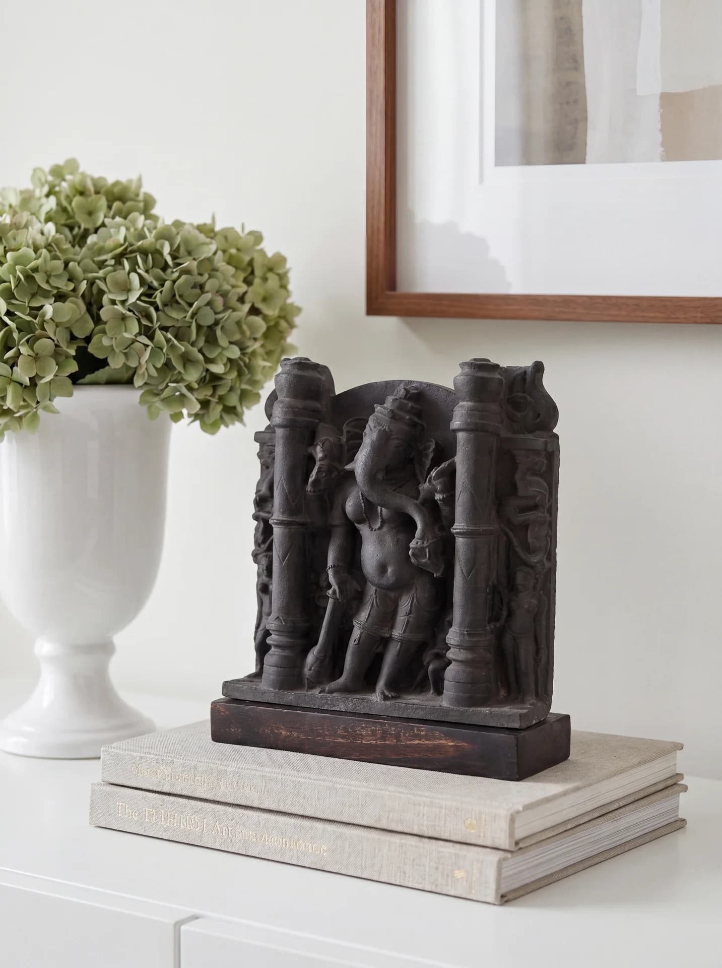 A dark stone Vinayaki Sculpture displayed on a stack of books next to a white vase of flowers.