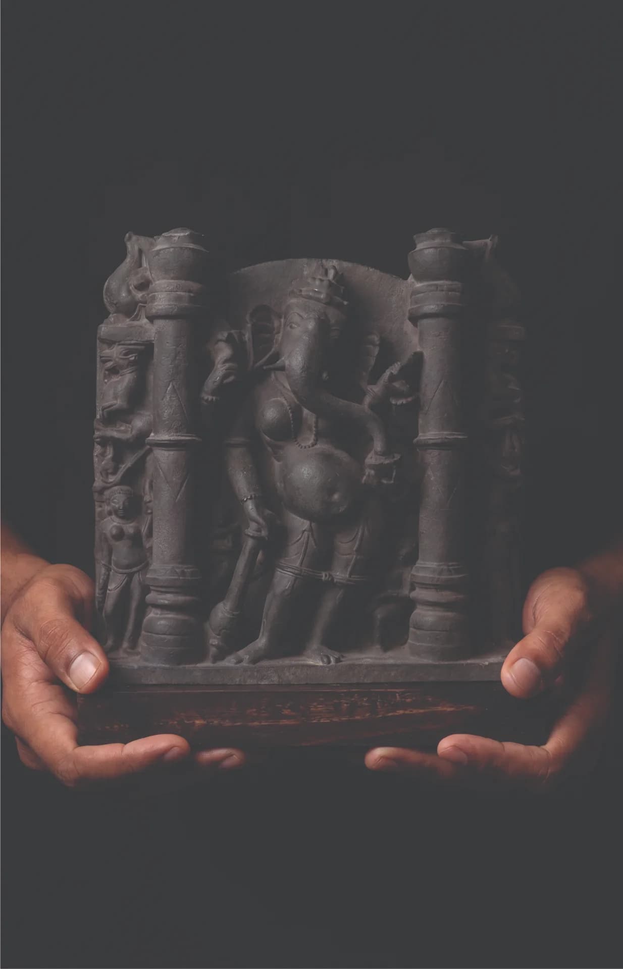 A pair of hands holds a dark stone Vinayaki Sculpture, a museum-grade replica featuring intricate temple art carvings.