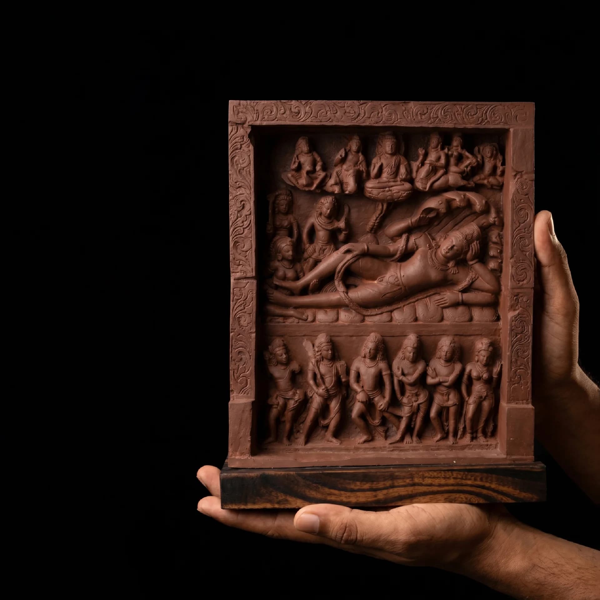Hands hold a terracotta-style Anantasayana Vishnu Sculpture, displaying the intricate relief of the reclining deity on a dark background.