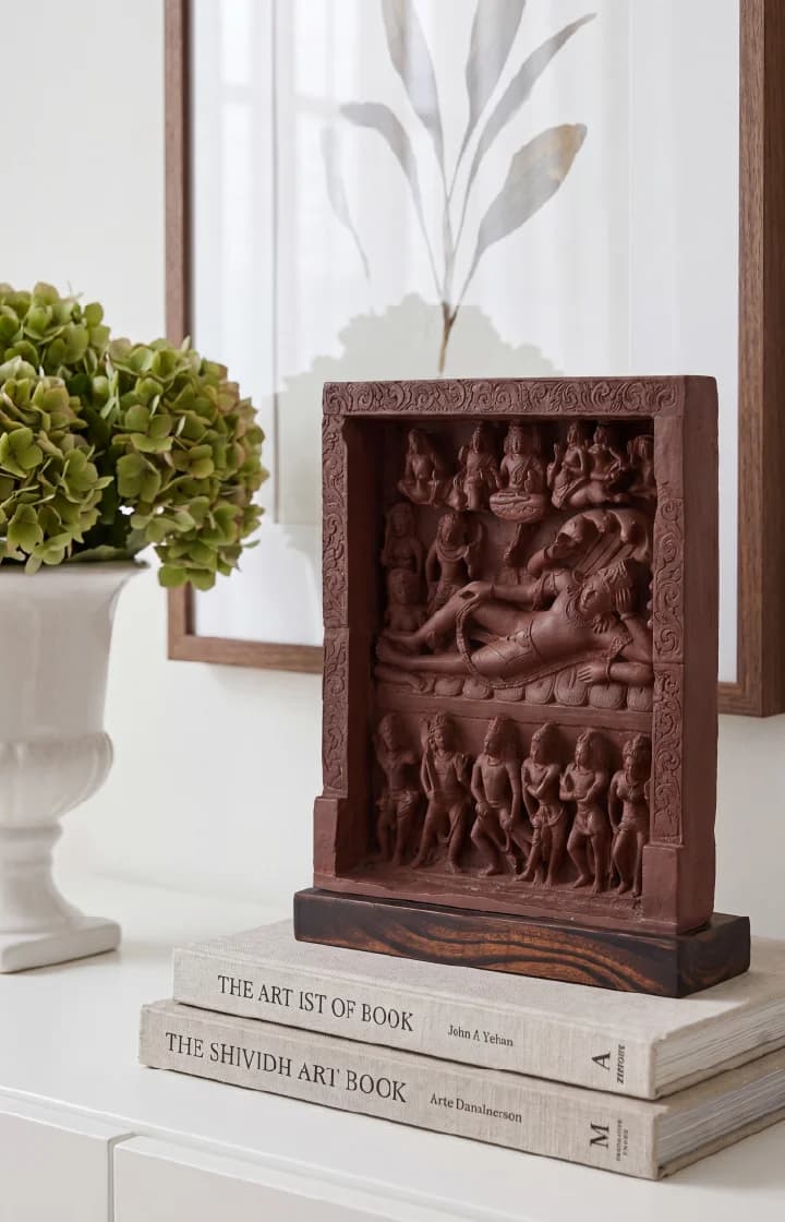 An Anantasayana Vishnu Sculpture replica in terracotta finish displayed on stacked books next to a white flower vase.