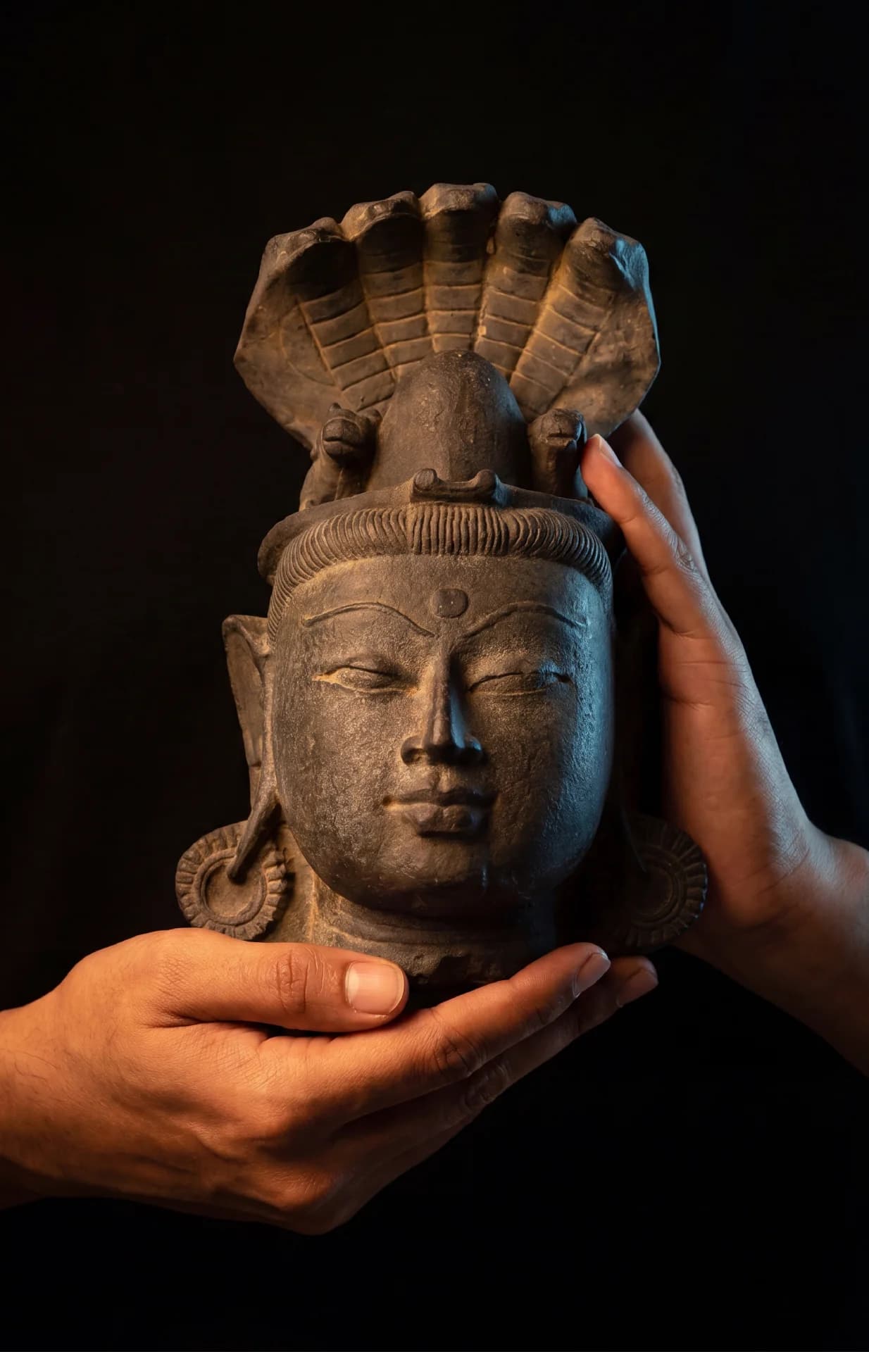 Two hands gently cradle the stone composite Shiva Lingam with Nandi & Serpent, highlighting the serene facial features.