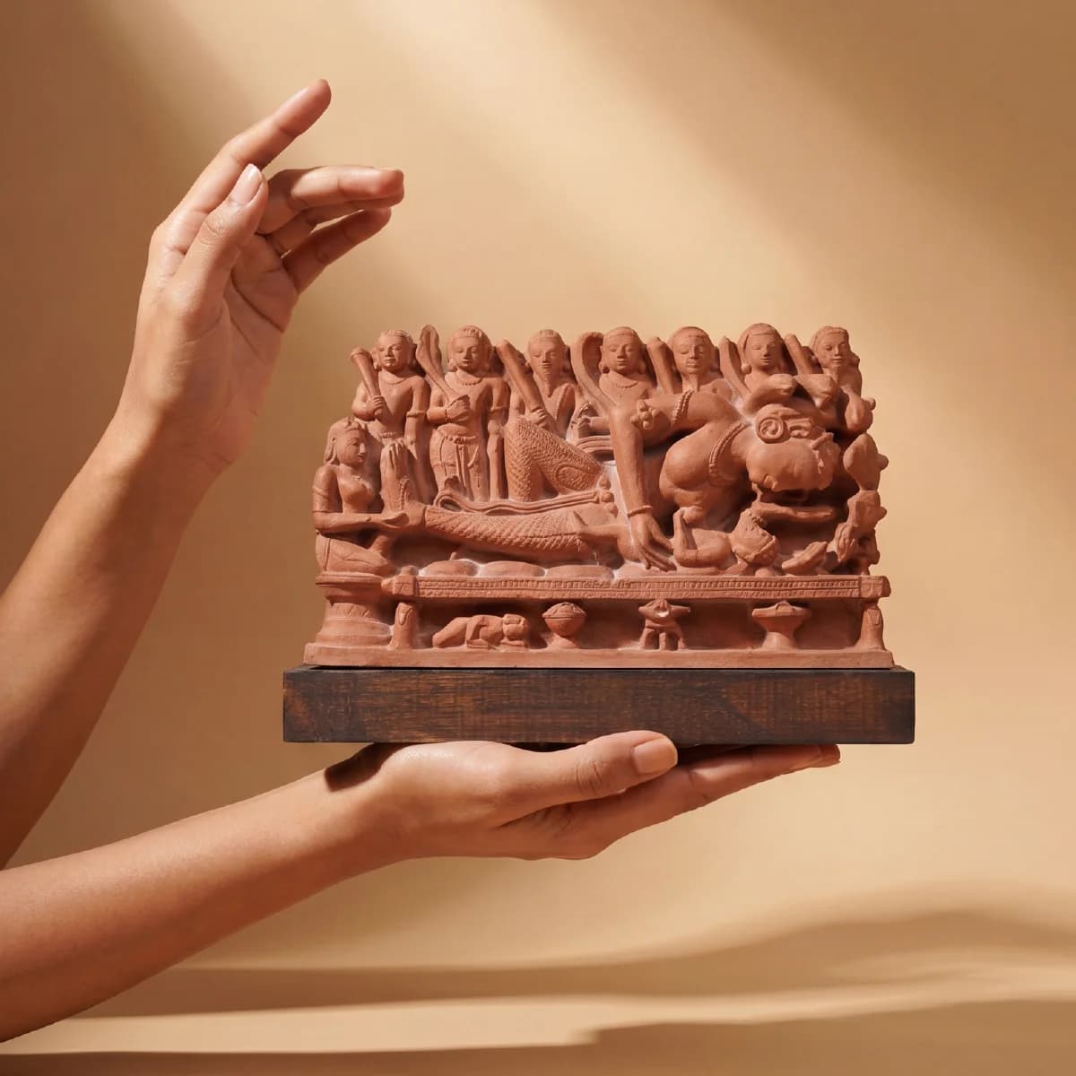 A pair of hands holds a handcrafted Krishna's Birth Sculpture, a detailed terracotta-style museum replica depicting ancient temple art.