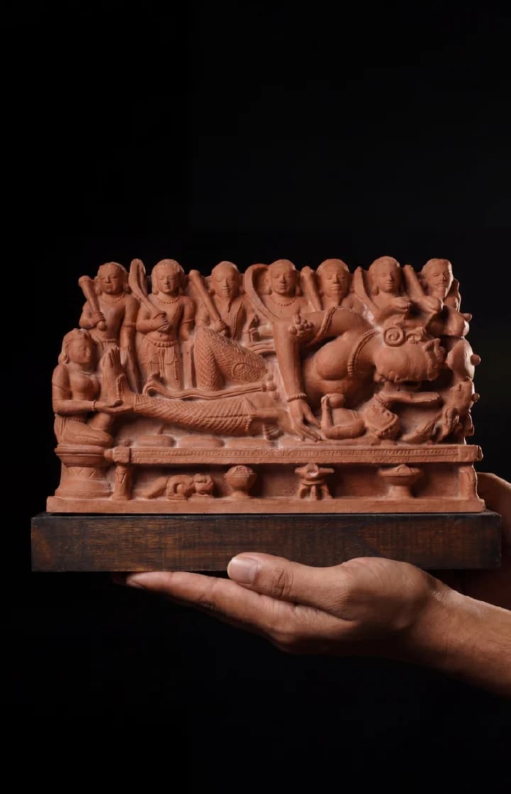 A person holds a terracotta-style Krishna's Birth Sculpture, showing intricate carvings of Devaki and guards against a dark background.