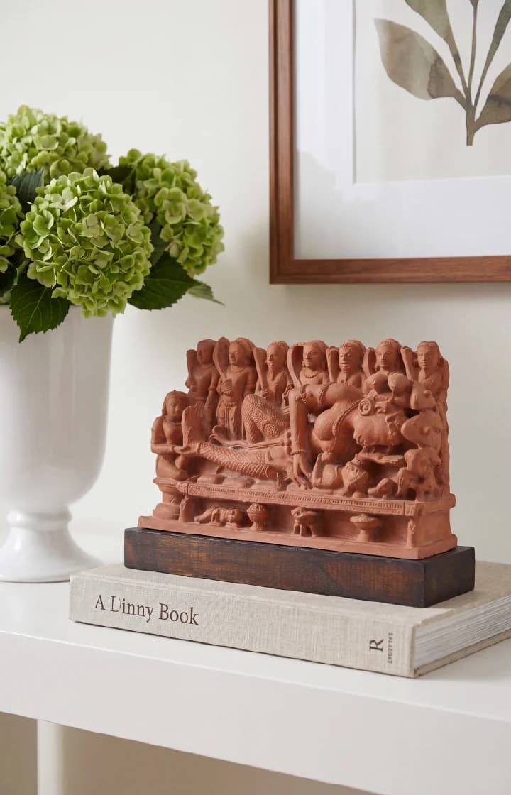 A terracotta-style Krishna's Birth Sculpture displayed on a book next to a vase of green flowers.