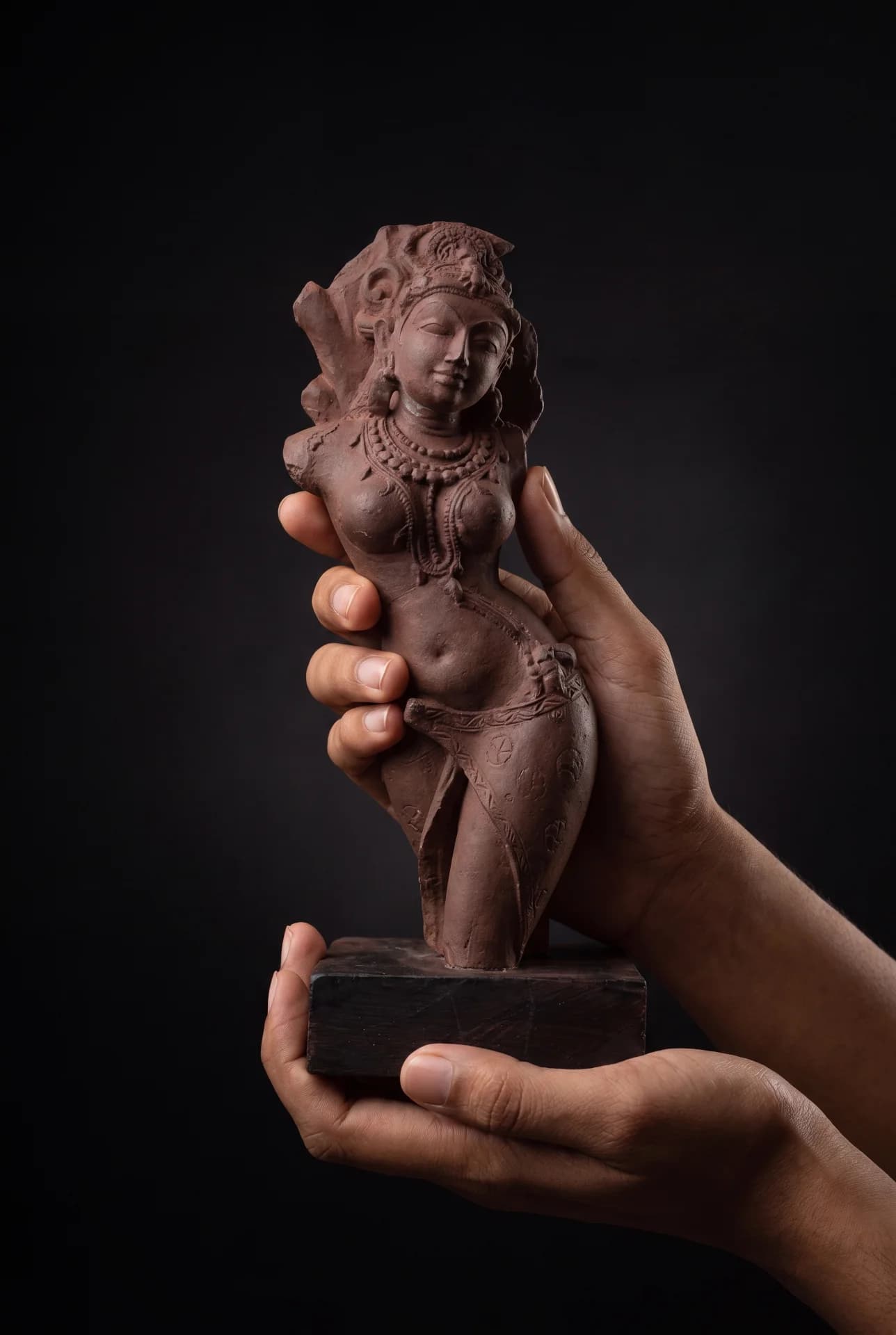A pair of hands gently holds a Salabhanjika Sculpture, showcasing the intricate stone composite detail against a dark background.