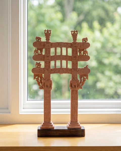 A Sanchi Torana Gateway Miniature crafted from stone composite, standing on a wooden base against a bright window.
