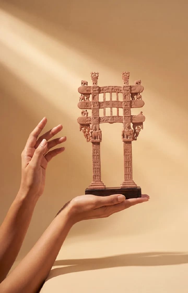 A pair of hands holds a stone composite Sanchi Torana Gateway Miniature against a soft, warm-toned studio background.